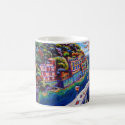 This artistic coffee mug shows a picture of colorful buildings on the side of a mountain at the base of the ocean.