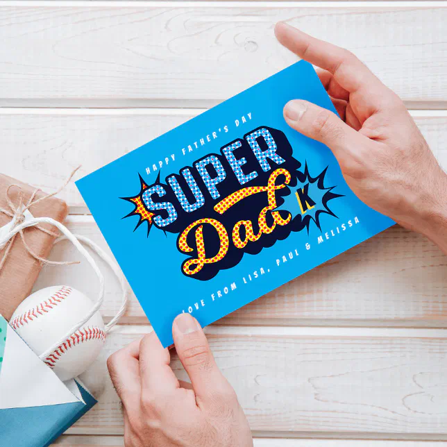 Two hands hold a blue superhero-themed Father's Day card. A baseball and other gifts can be seen to the left.