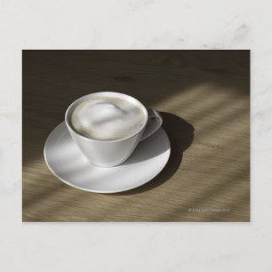 A cup of cappuccino coffee lies on an oak postcard