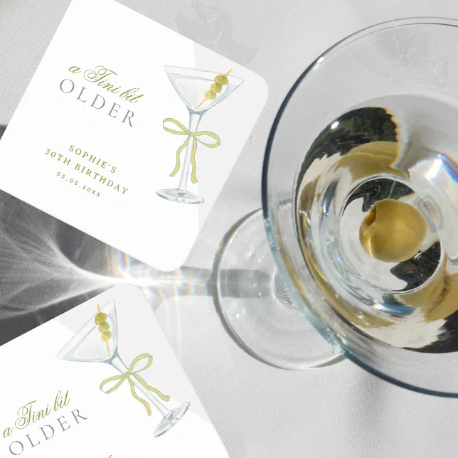 Two coasters with a martini design are shown lying on a table. A drink in a martini glass with an olive in it is on the right.