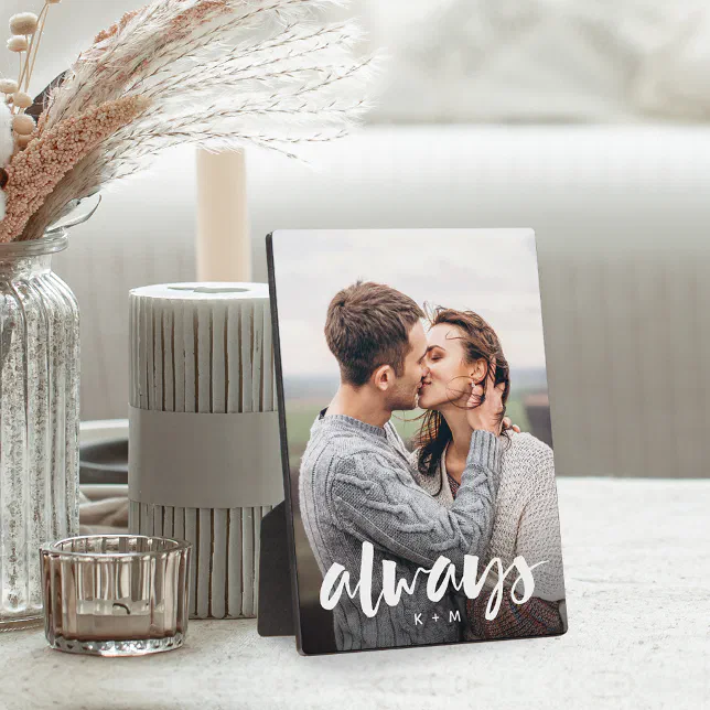 A photo plaque pictured resting on a table with dried flowers and a candle to the left, with a blurred white couch in the background.