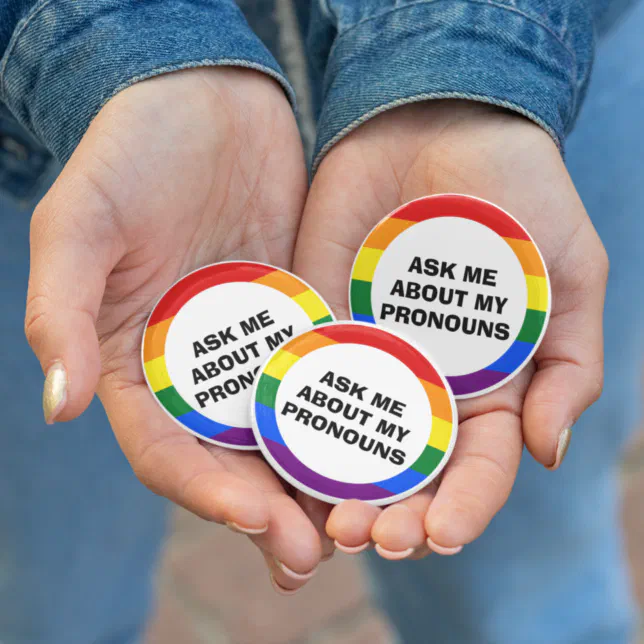 Three pronoun-focused buttons are pictured in the palms of a person hands. 