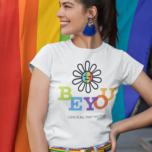 A person is smiling wearing a Pride-themed t-shirt, standing in front of a Pride flag. 