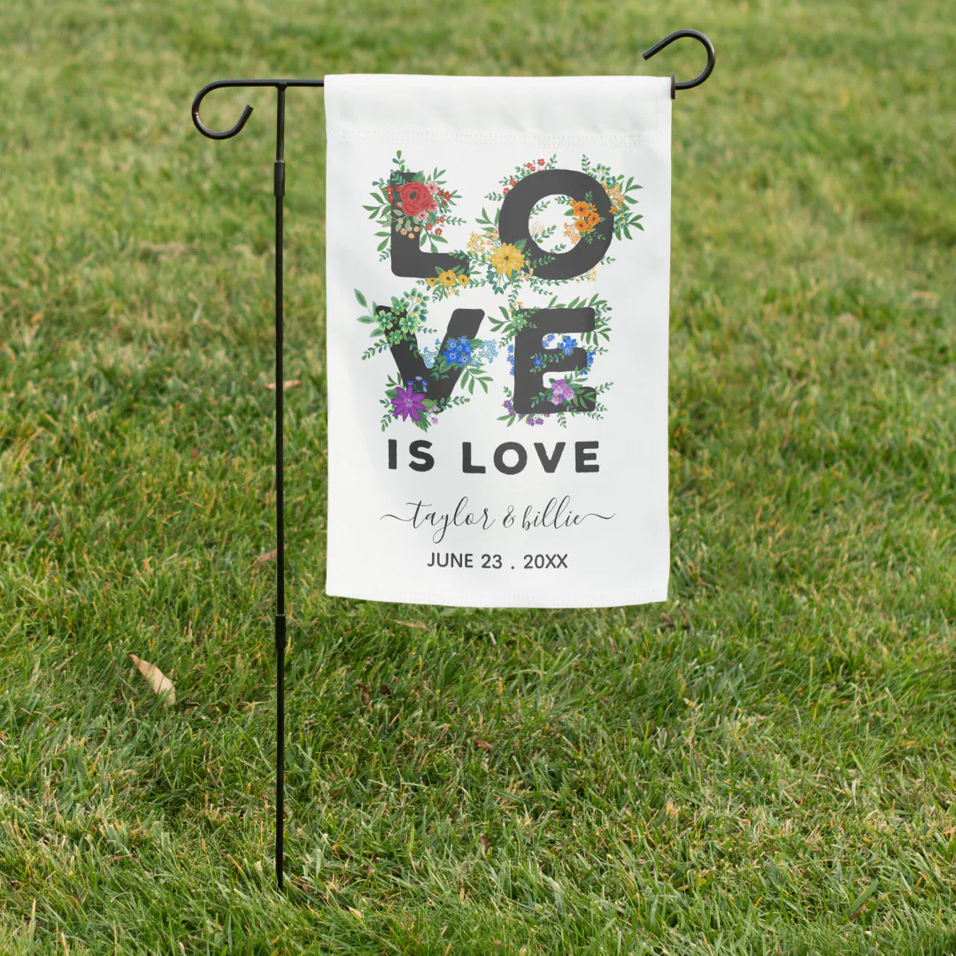 A white garden flag on a black metal stake, set in a grass lawn, decorated with large bold letters intertwined with colorful wildflowers and greenery in red, yellow, blue, and purple.