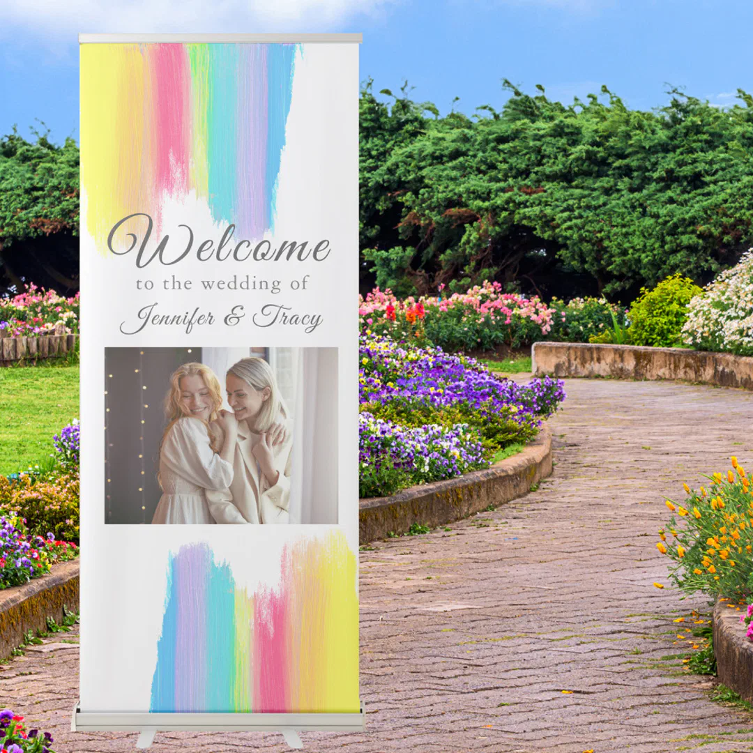 A tall retractable banner with a white background and soft pastel rainbow paint brush strokes, featuring a photo of two women in cream outfits laughing together, displayed outdoors on a garden path surrounded by colorful flower beds.