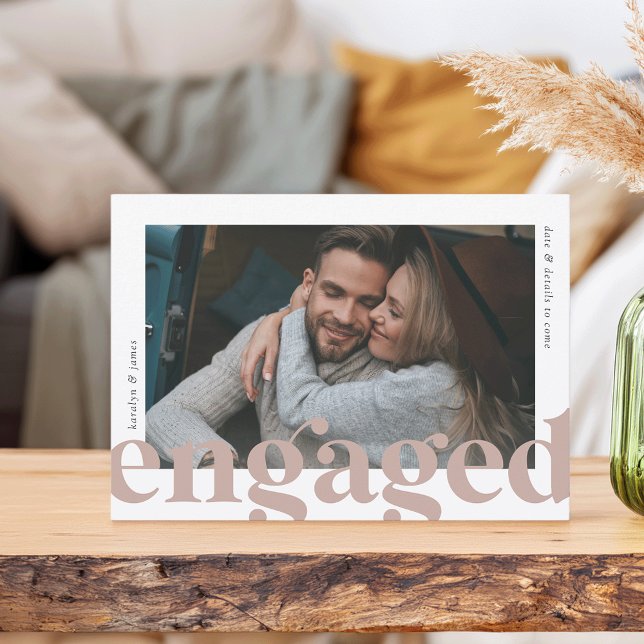 A photo engagement anouncement card. A woman is shown hugging her fiancé. The card is propped up on a wooden counter and the contents of a living room can be seen in the background.