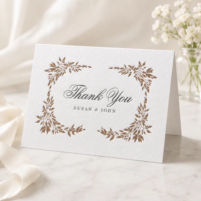 A copper-colored thank you card with a floral border. A bunch of white flowers in a vase can be seen to the right.