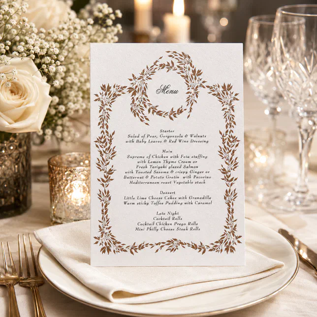 A copper-colored wedding menu with a floral wreath and border. There are flowers and glasses in the background, and it is standing on a cloth napkin.