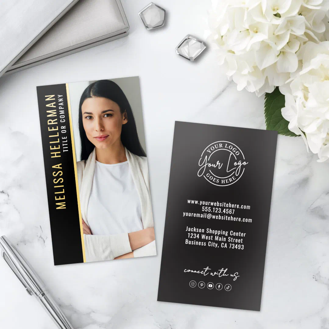 The front and back of the business card are on display which includes a photo option, white and gold text, on a black background. The card itself is on a white marble mantle with white flowers to the right.