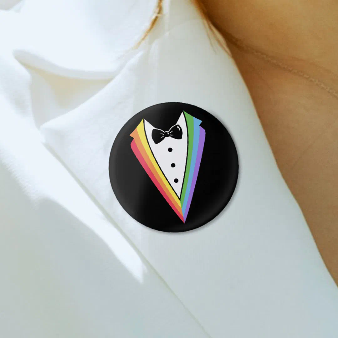A round black pin badge featuring an illustrated tuxedo with rainbow-colored lapels and a bow tie, pinned to a white shirt collar.