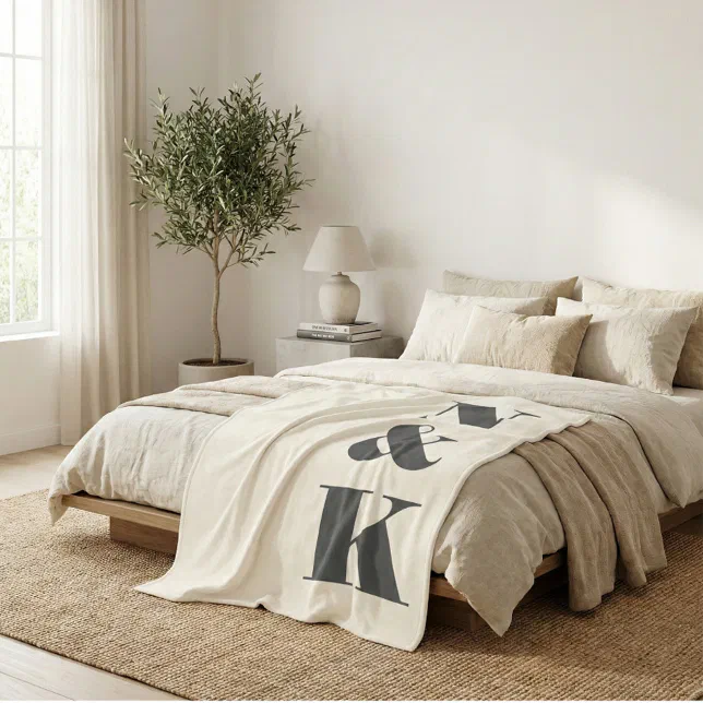 A bed with cream and beige colored pillows and an printed throw blanket with custom initials. In the background there is a large green potted plant and a side table with a white lamp.