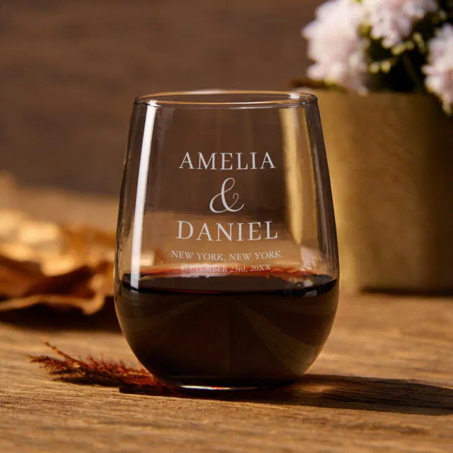 A stemless wine glass positioned on top of a brown, wooden table. It is filled about a third of the way with red wine and behind it is a flower pot containing pink flowers.