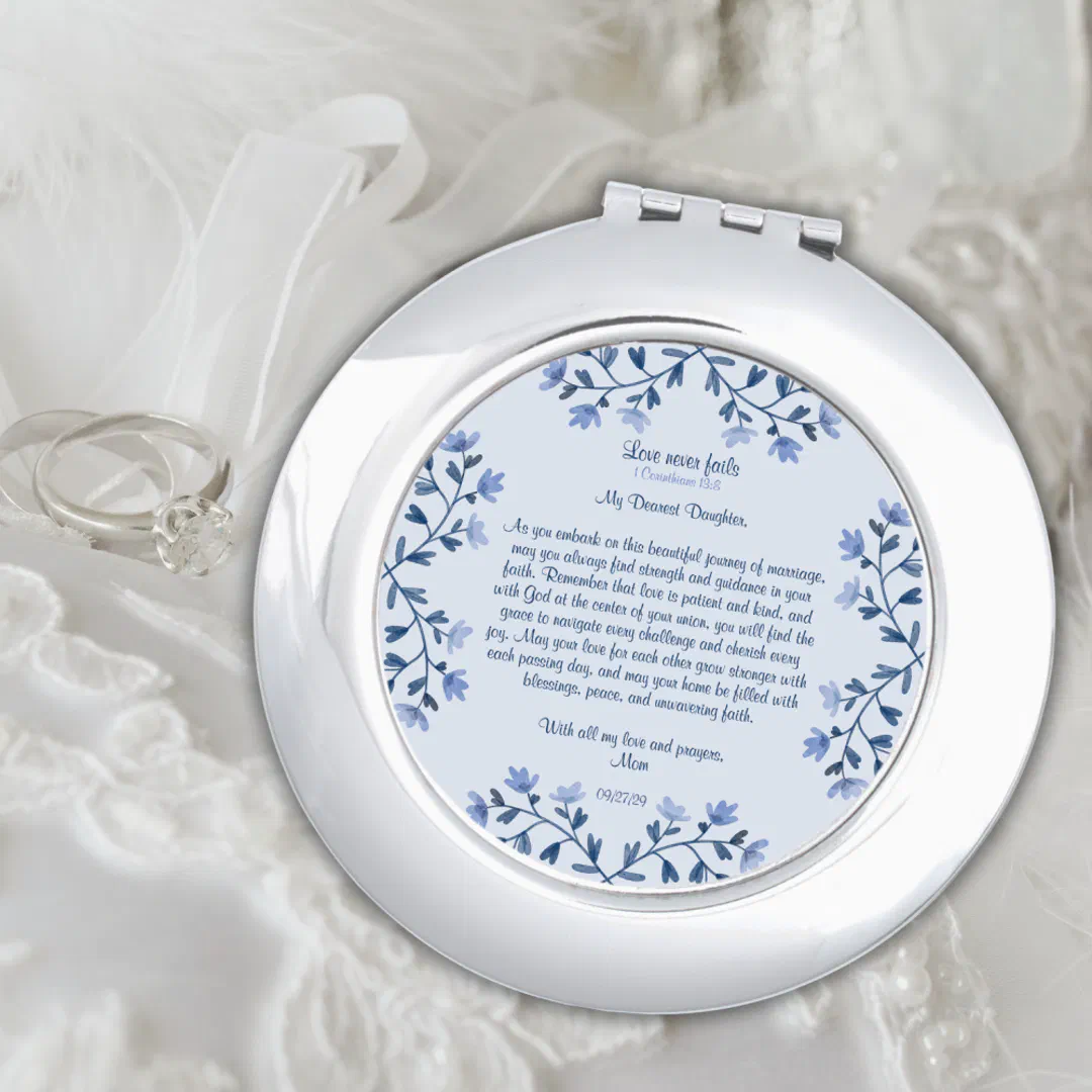 A personalised round compact mirror with a dusty blue floral cover sitting on white satin next to a dress ring.