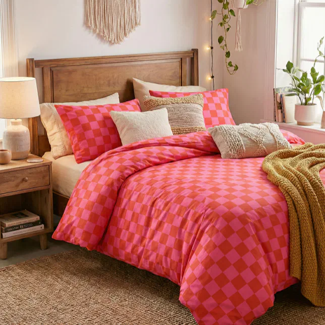 A brown wooden bed with pink and orange checkered sheets and cream and beige pillows. In the background there is some houseplants and a wooden sidetable with a cream lamp.
