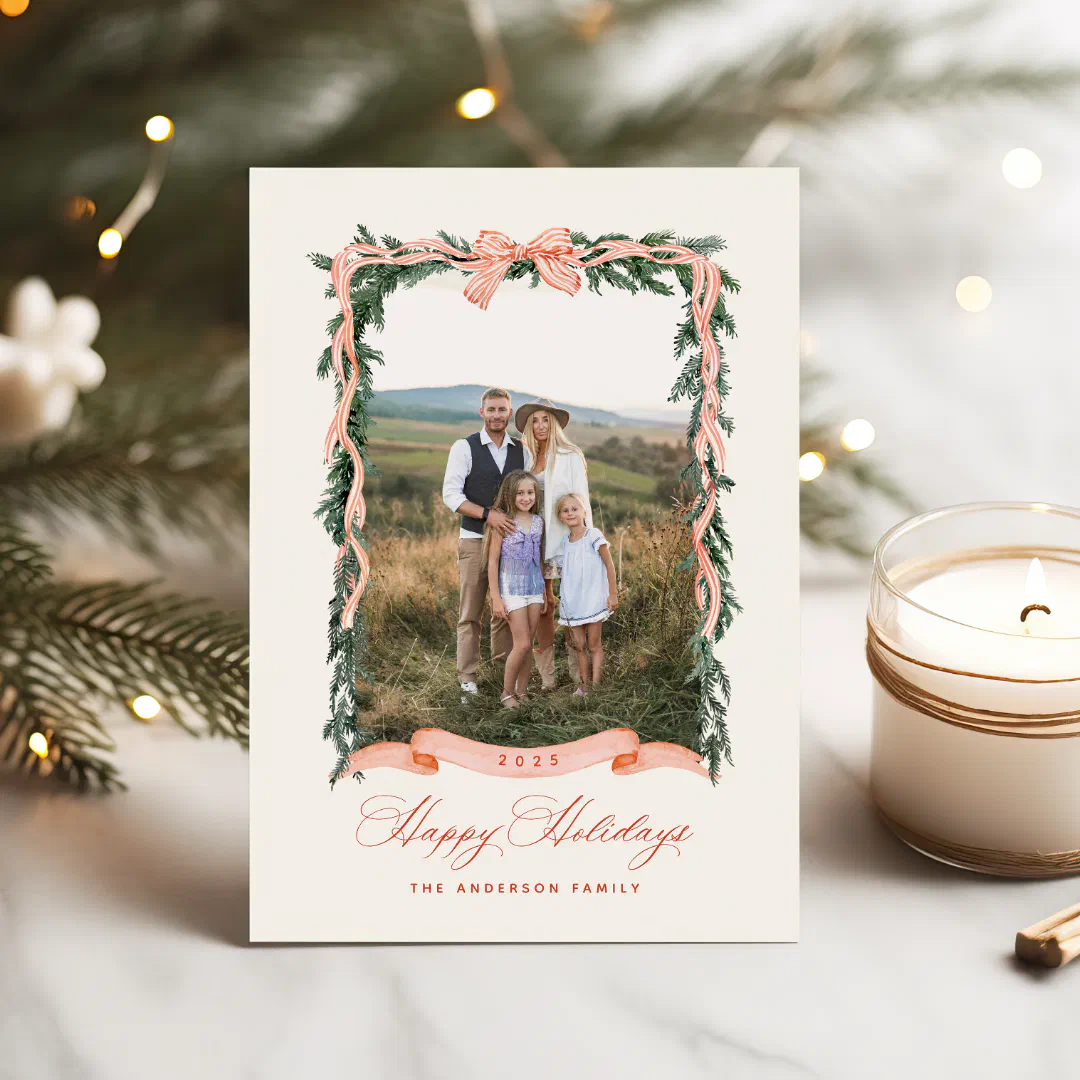 A greenery wreath and bow style holiday card is shown next to a white candle with a Christmas tree in the background.