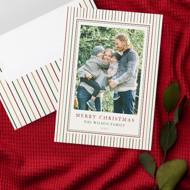 A red, green and gold striped envelope placed next to matching a striped holiday card on a red blanket with some leaves in the background.