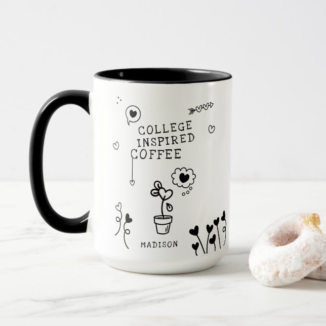 A coffee mug with with some designs and some writing that says "college inspired coffee". It is on a marble countertop with a donut beside it and a plain background.