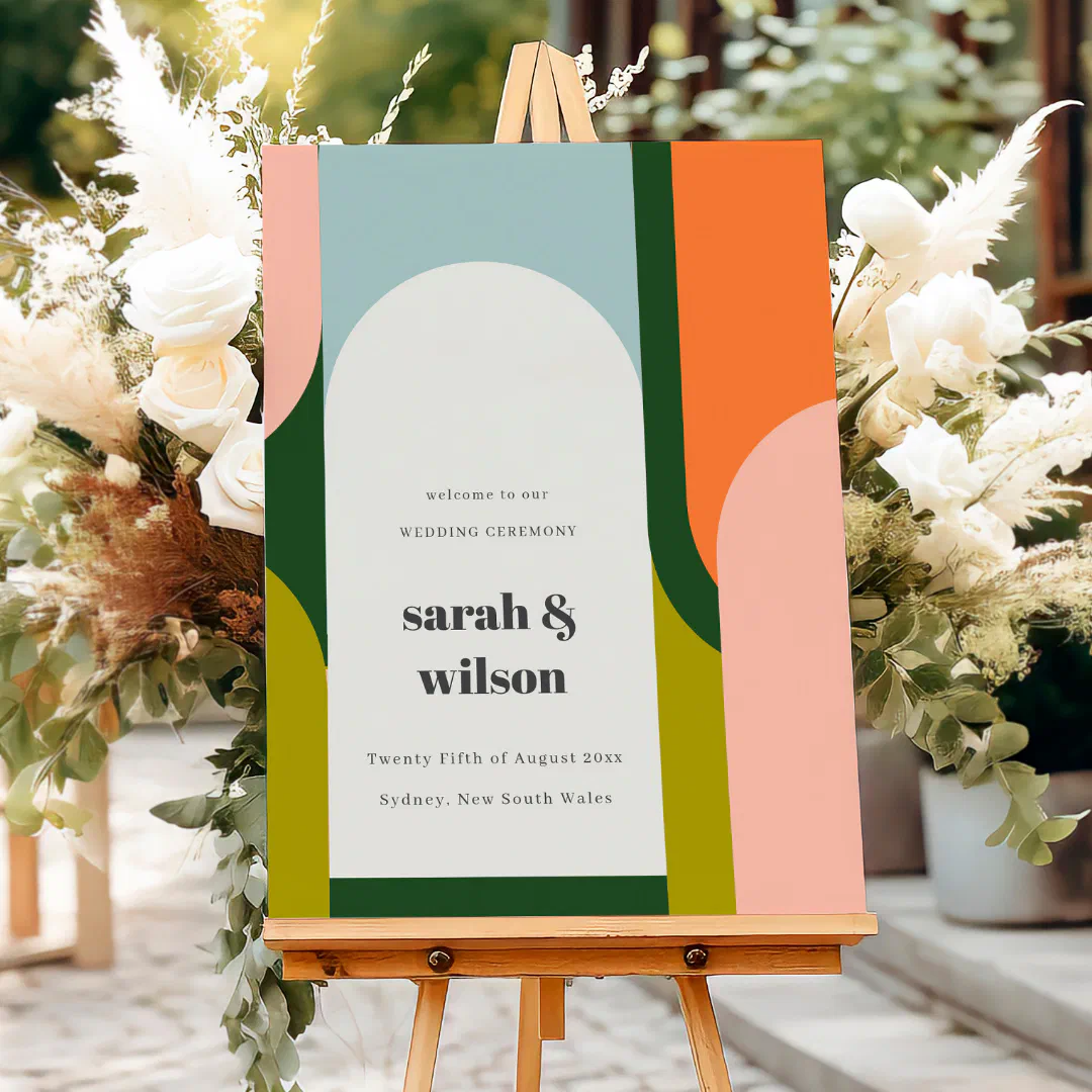A colorful groovy wedding welcome sign with bold retro arches in orange, pink, green, and blue, displayed on a wooden easel flanked by white roses, pampas grass, and greenery on a sunlit courtyard path.
