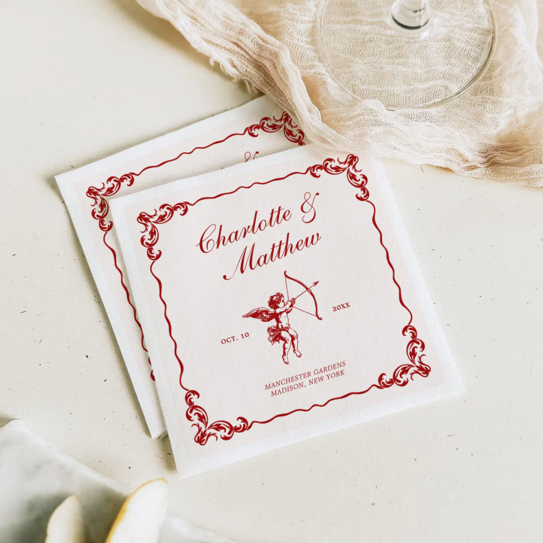 Vintage-style wedding napkins with a red cherub illustration and ornate scrolling border, placed on a cream table beside a wine glass and gauzy cloth.