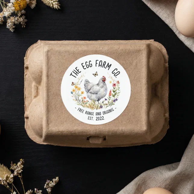 A sticker is seen on top of a carton of eggs, sitting atop a black mat, with an eggs and dried flowers seen in the corner of the image.