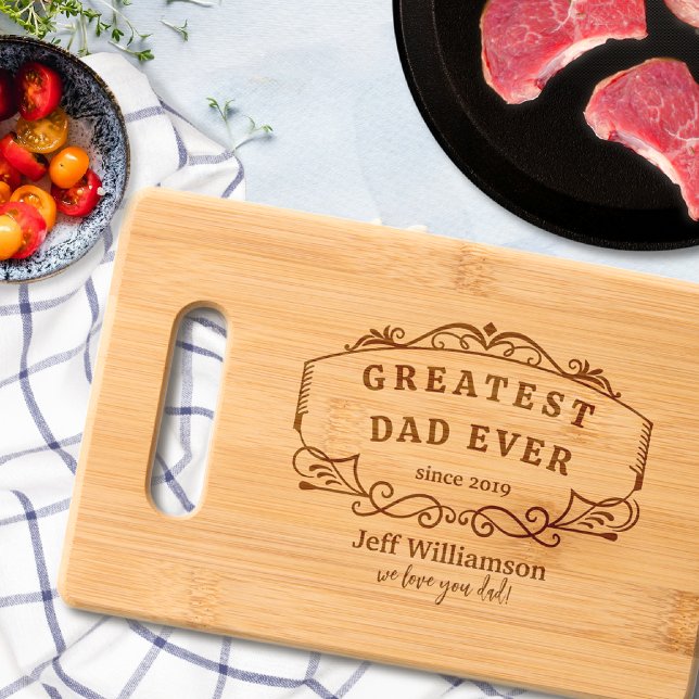 A brown cutting board with "Greatest Dad Ever" printed on the front, with a plate of meat and a bowl of tomatoes featured around it.