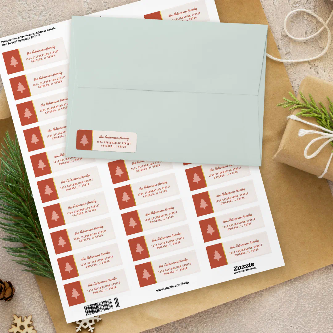 A sheet of return address labels with a pink Christmas tree on a red block, partly covered by a pale mint envelope with one label applied, on kraft paper with pine sprigs, twine, a wrapped gift, and wooden snowflakes.