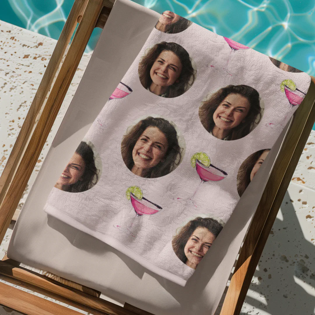 A light pink beach towel draped over a wooden sun lounger beside a sparkling turquoise pool, printed with a repeating pattern of a smiling woman's face in circular cutouts interspersed with illustrated pink cocktail glasses garnished with lime wedges.