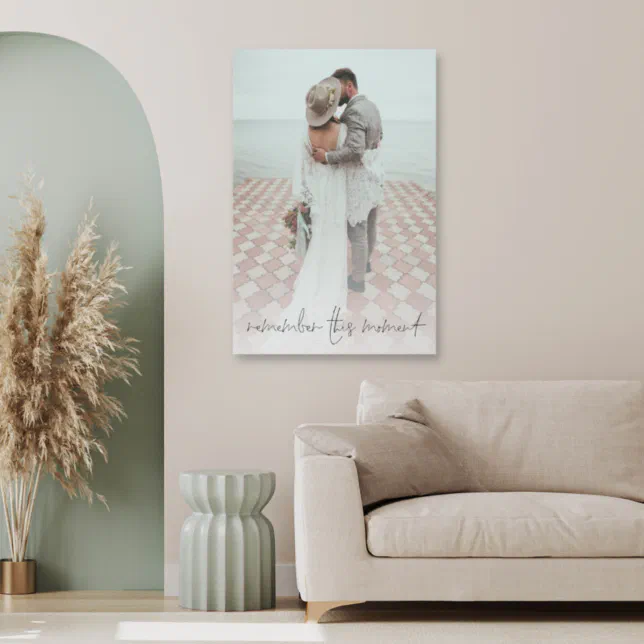 A photo print is place on a wall above a beige couch, with a green vase underneath and a plant to the left. 