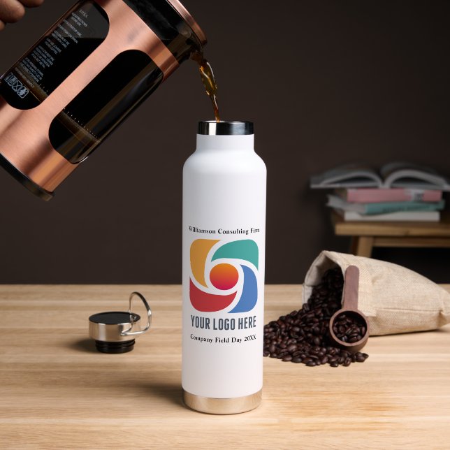 A white metal bottle is seen on a light brown table with coffee being poured into it. The cap of the bottle and coffee beans are seen in the background.