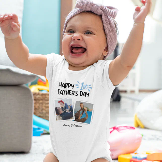 An excited baby is wearing a white body suit that says "Happy 1st Father's Day." There's some toys and a gray couch in the background.
