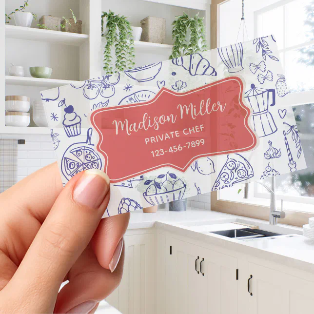 A person holds a clear business card with a cookery-themed design and a red emblem in the middle. A kitchen can be seen in the background.