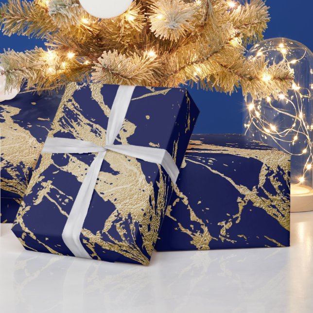 Gifts wrapped in navy and gold wrapping paper on a marble counter with gold Christmas branches and fairy lights placed in a glass jar in the background.