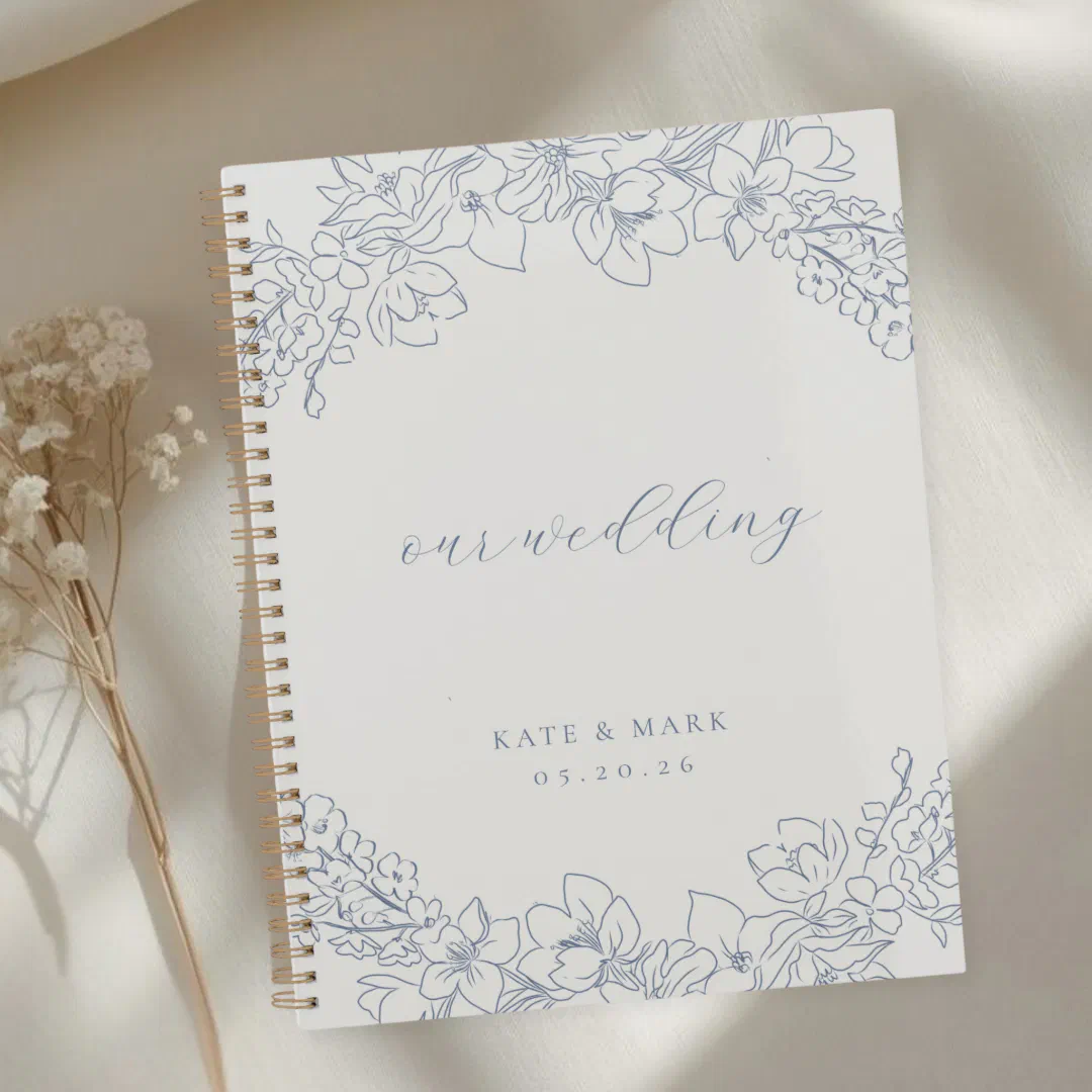 A hard cover spiral notebook with a delicate blue floral cover on a white background placed next to flowers.