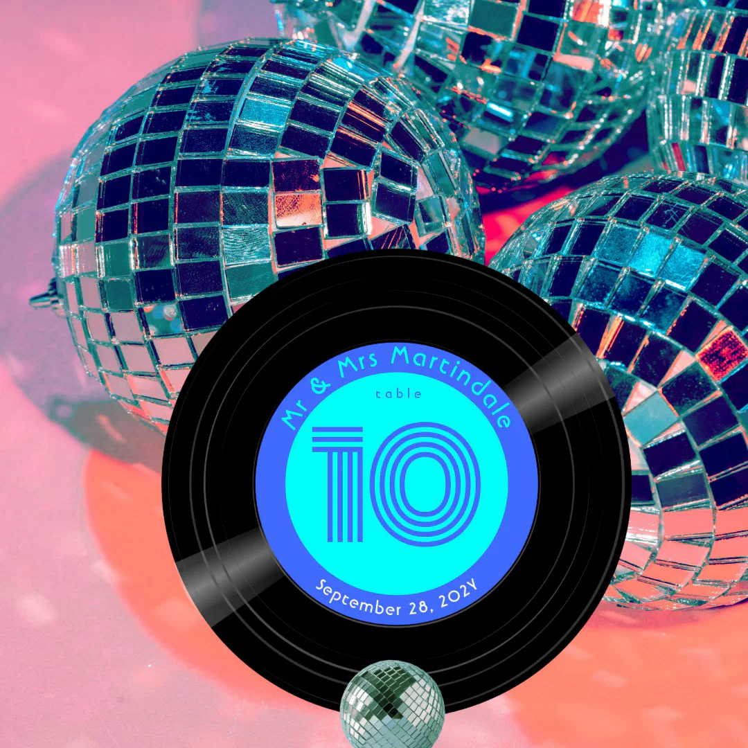 A wedding table number designed as a vinyl record with a bright blue label, set against shimmering disco balls in pink and blue lighting.