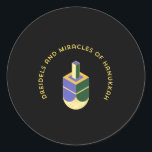 DREIDELS AND MIRACLES OF HANUKKAH CLASSIC ROUND STICKER<br><div class="desc">This stylized illustration features a dreidel colored yellow, purple, and green against a stark black background. A curved gold text above it reads "DREIDELS AND MIRACLES OF HANUKKAH", highlighting the traditional toy and the spiritual celebration of the Jewish holiday. The design has a clean, geometric aesthetic with a touch of...</div>