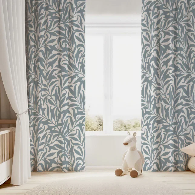 White and dusty blue leafy printed curtains shown on a large window. In the background there is a baby crib and a stuffed animal. 