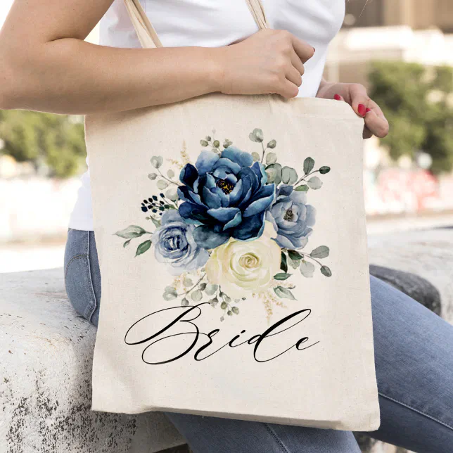 A canvas tote bag with blue and white roses on it and held by a woman in a t-shirt and jeans.