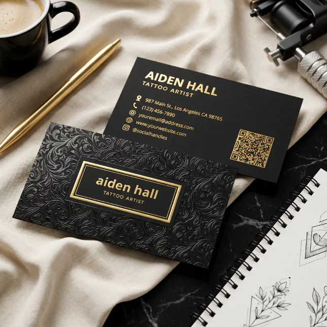 A black and gold business card featuring a QR code is lying next to a notebook, pen, and mug.