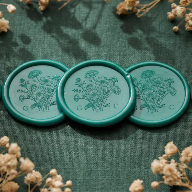 A set of three green wax seals with a flower motif. They are lying on a textured green material with foliage in the background.