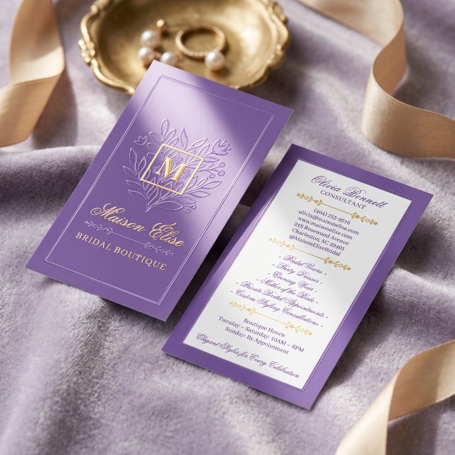 An elegant, double-sided rectangular business card with a gold foil crest and border sits on a textured lavender fabric backdrop alongside gold silk ribbons and a decorative gold jewelry dish holding a pearl necklace.