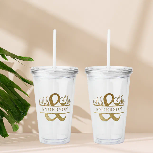 Two custom clear drink tumblers with printed glittery gold lettering. In the background there is a green plant.


