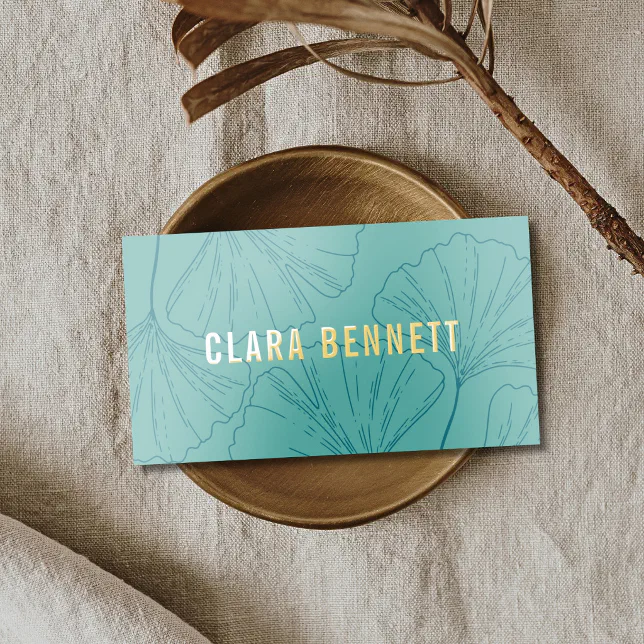 A chic mint green business card with a delicate ginkgo leaf line art pattern and gold foil typography resting in a small brass dish on a textured linen surface.
