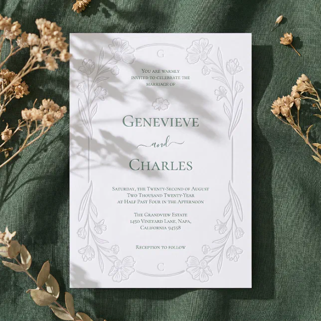 A green letterpressed wedding invitation lying on a textured green material with foliage in the background.