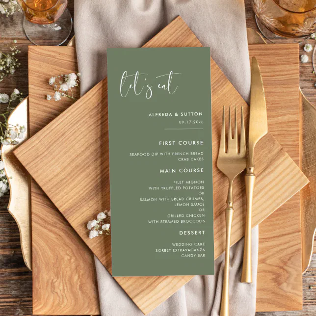A green-colored wedding menu. The menu is sat next to a knife and fork and is resting on top of serving boards. Drinkware can be seen in the top-left and top-right corners.