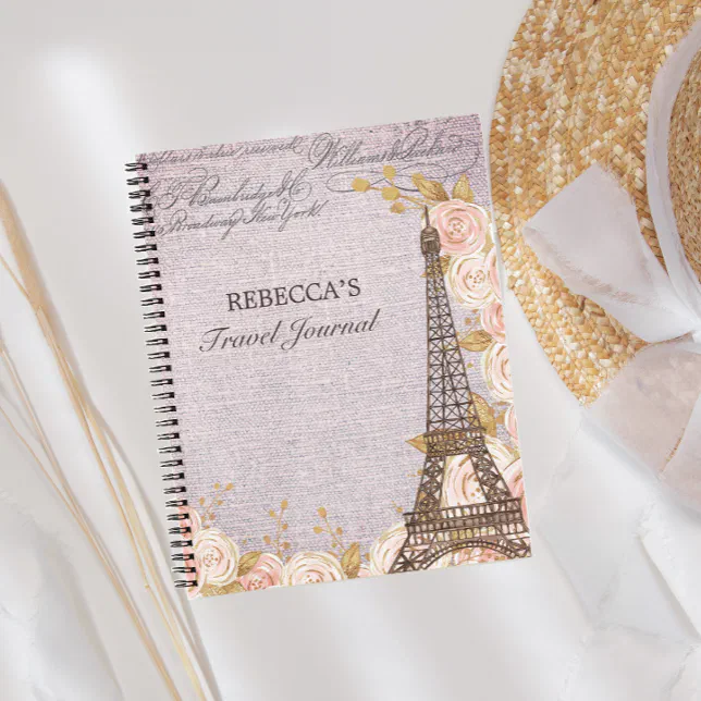 A Paris-themed custom travel journal with a pink and white rose design pictured next to a sunhat with a white bow tied around it.