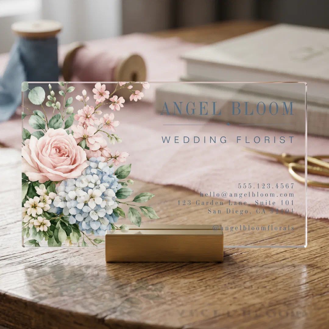 A transparent business card with a flower bouquet on the left with gray text. It's placed on a wooden card holder with a sewing kit and books in the background.