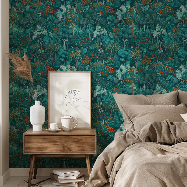 A brown wooden table sits next to a bed with beige bedding. On the table there is a white mug and a white vase as well as a framed piece of artwork with a simple drawn hand. On the wall in the background there is a teal and green forest-style wallpaper.