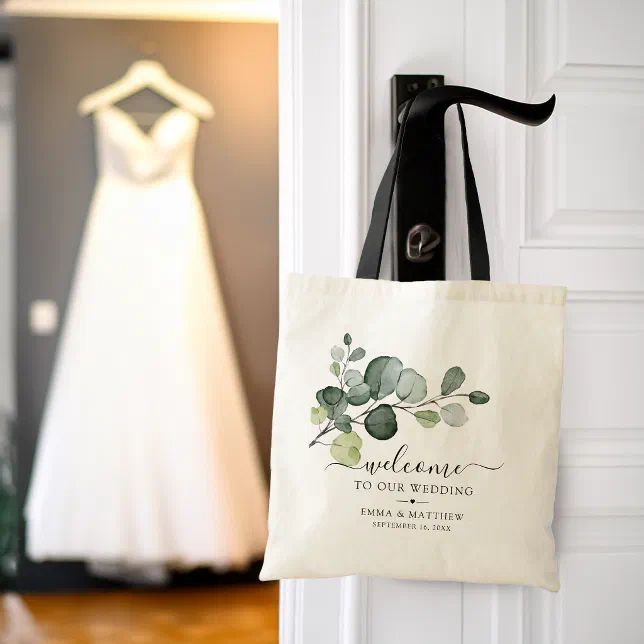 A tote bag welcoming guests to a wedding is hanging from a door. A wedding dress can be seen hanging in the background.