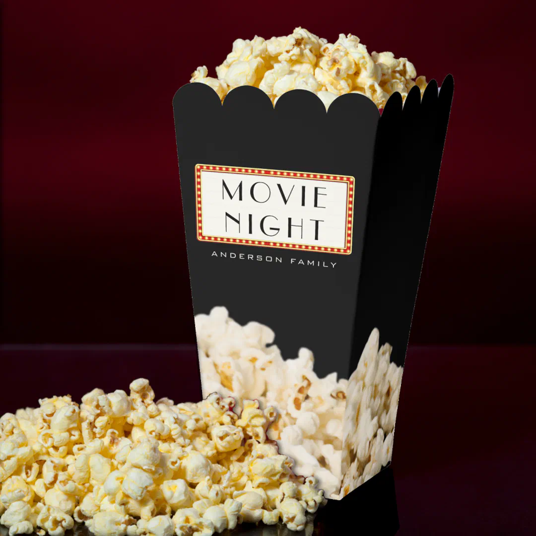 Black popcorn favor box with a theater marquee sign design personalized with a family name, overflowing with popcorn.