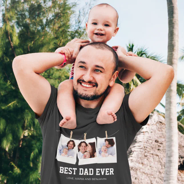 A father is wearing a black t-shirt that says "Best Dad Ever" with his baby on his shoulders. There's a blue sky and trees in the background.